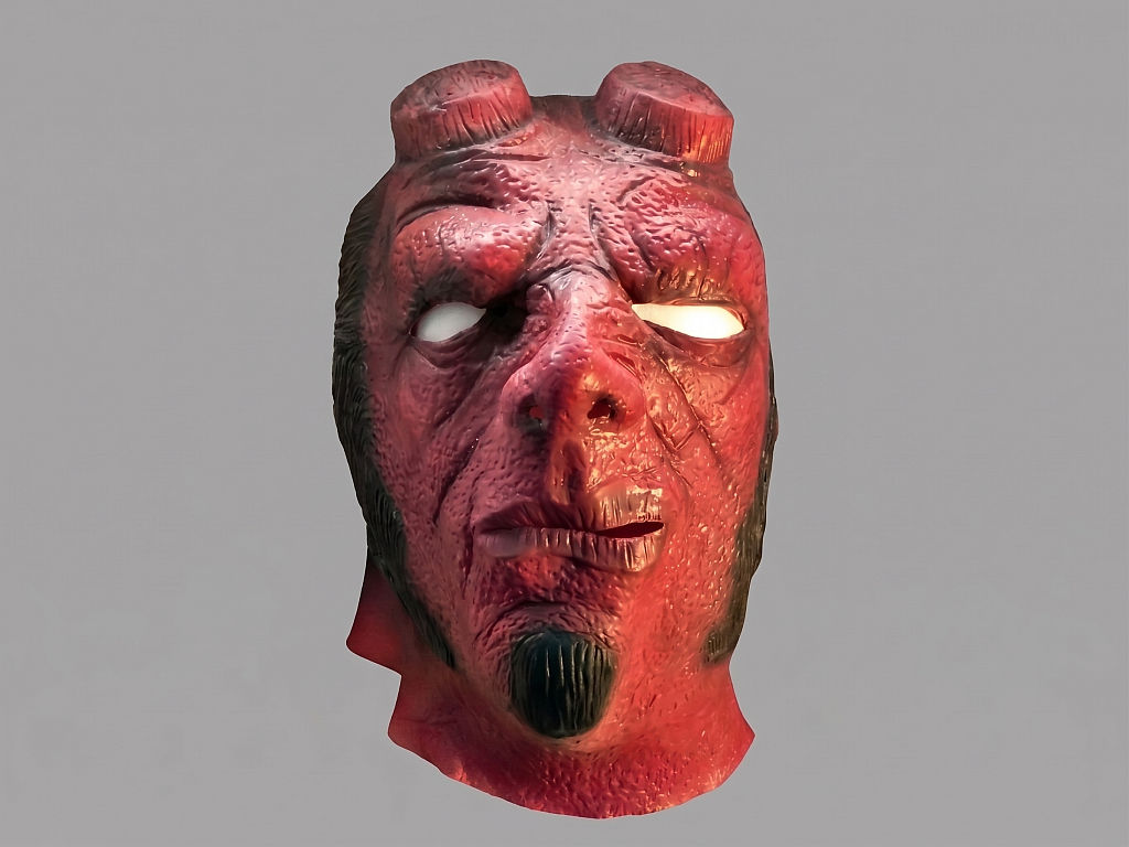 Groteske, verwitterte rötlich-braune Höllenmonstermaske mit angebrochenen Hörnern, Frontalansicht vor grauem Hintergrund.